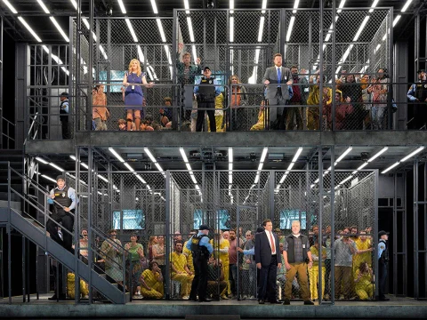 A two-story indoor jail scene with multiple fenced cells, guards in blue uniforms, and inmates. Some inmates are in yellow jumpsuits, while others wear casual attire.