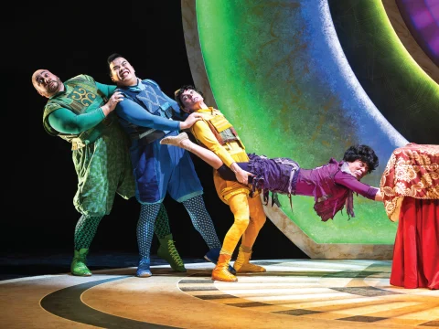 Four actors in colorful costumes perform a humorous, exaggerated pose on stage against a vibrant, circular backdrop.