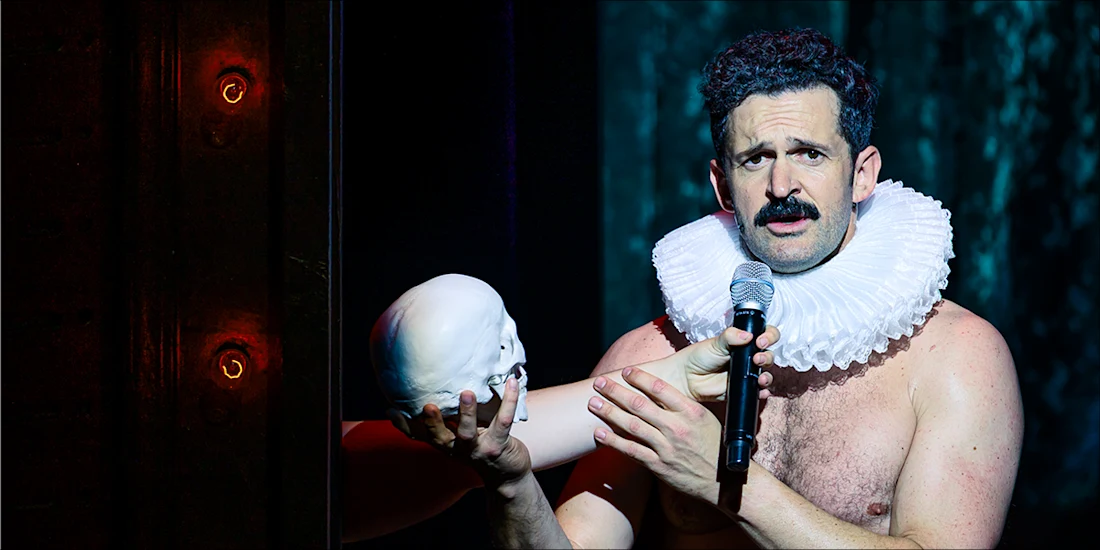 A man with a ruffled collar holds a microphone and a skull while performing on stage under dramatic lighting.