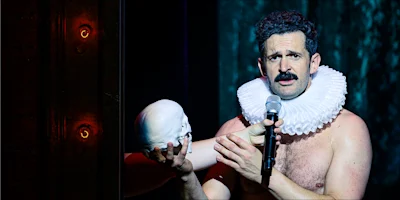 A man with a ruffled collar holds a microphone and a skull while performing on stage under dramatic lighting.