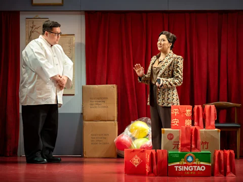 A man in a white chef coat and a woman in a patterned blazer converse on stage. Boxes, balloons, and red gift bags are stacked nearby in front of a red curtain.