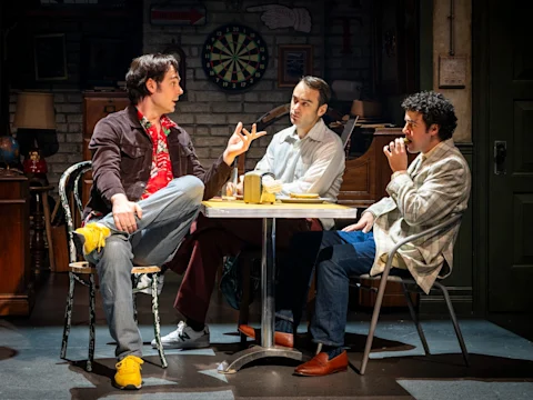 Three men sit around a small table in a dimly lit room, engaged in conversation, with a dartboard and various objects visible in the background.