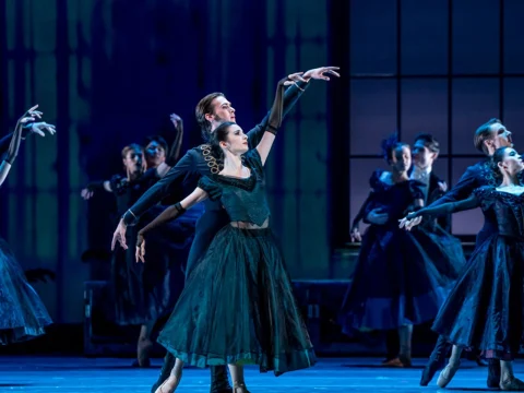 Dancers in dark costumes perform a ballet on stage, with two main performers in the foreground and several other dancers in the background. The setting is dimly lit with a window-like structure.