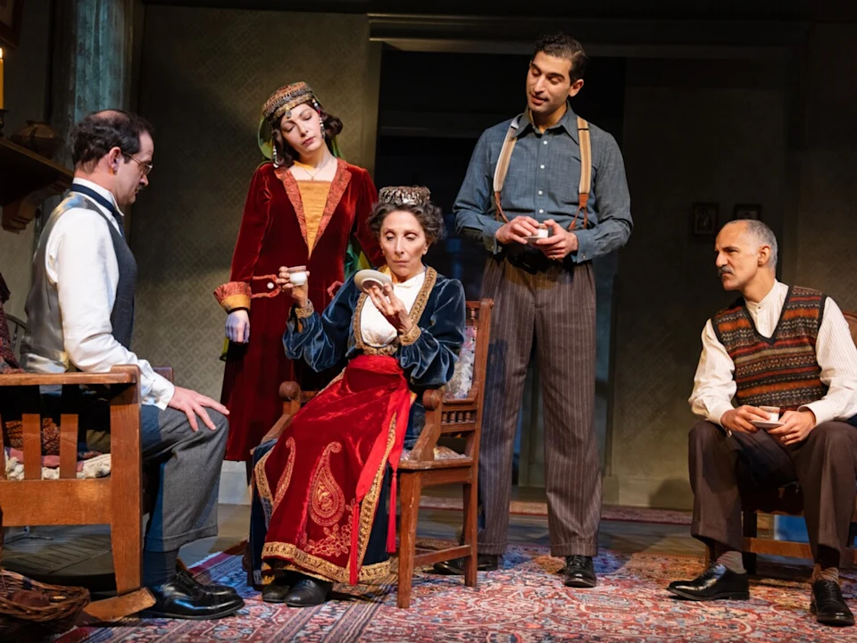 Five actors in period costumes perform a scene on stage, with one woman seated and the others standing or sitting around her in a domestic setting.