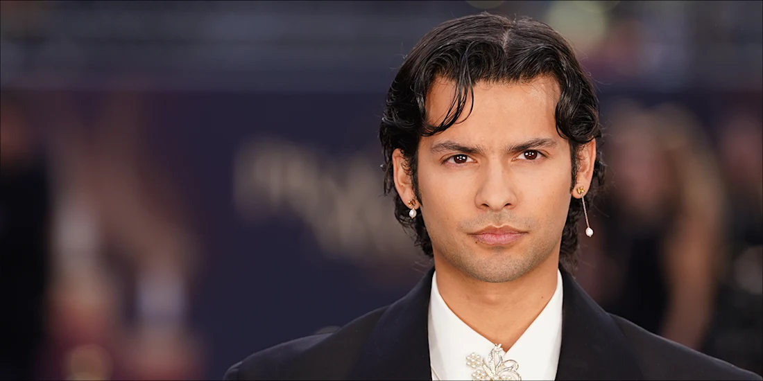 A person with dark, wavy hair and earrings, wearing a black suit jacket and white shirt, looks directly at the camera against a blurred background.