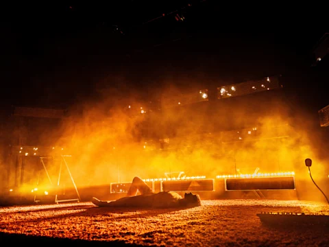 A person lies on a carpeted floor in a dimly lit space, surrounded by orange and yellow stage lights and smoke effects.