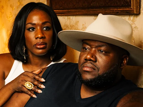 A woman with shoulder-length hair rests her hand on the shoulder of a bearded man wearing a wide-brimmed hat. Both have serious expressions and are indoors with patterned wallpaper.