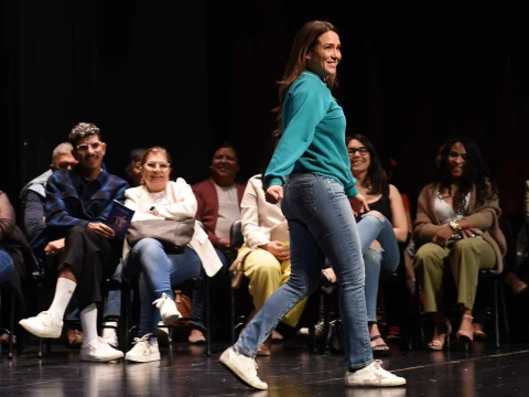 A woman in a teal sweater and jeans walks confidently on stage while an audience watches, smiling and casually dressed.