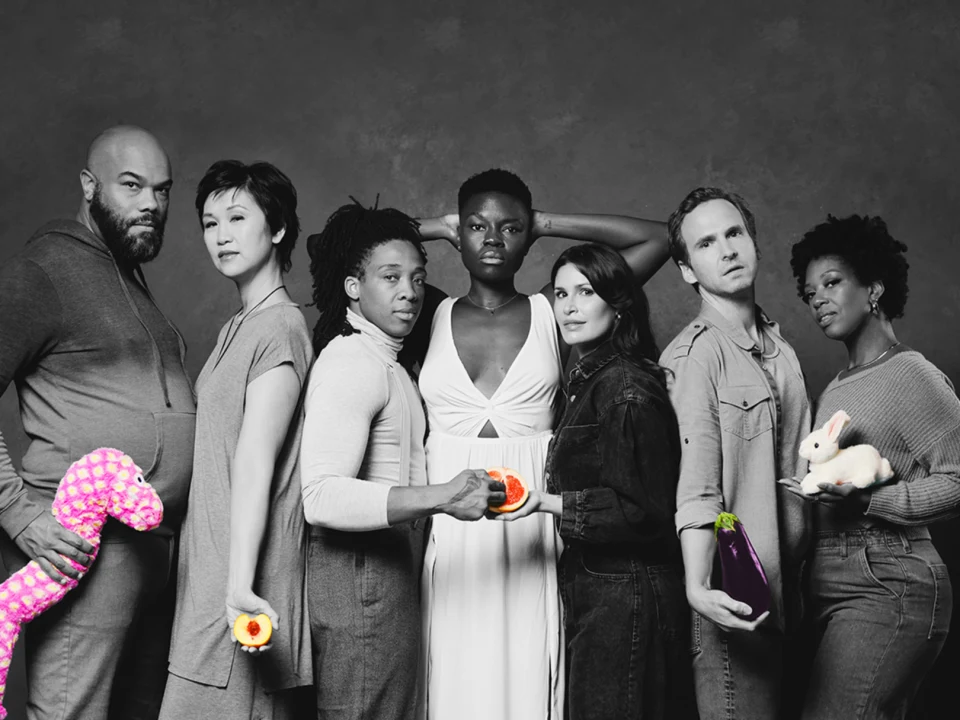 Cast standing in black and white holding colorful objects like a bunny, grapefruit, flower, and stuffed animal.
