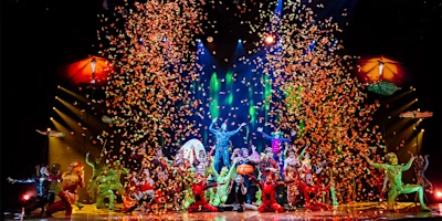 A colorful stage performance with multiple costumed performers, dramatic lighting, and a shower of orange confetti falling from above.