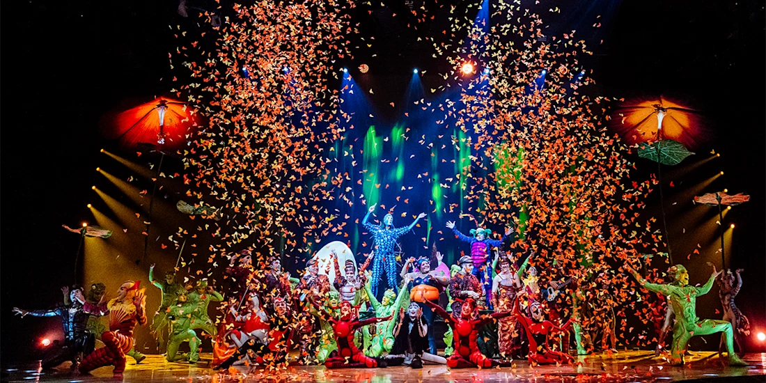 A colorful stage performance with multiple costumed performers, dramatic lighting, and a shower of orange confetti falling from above.