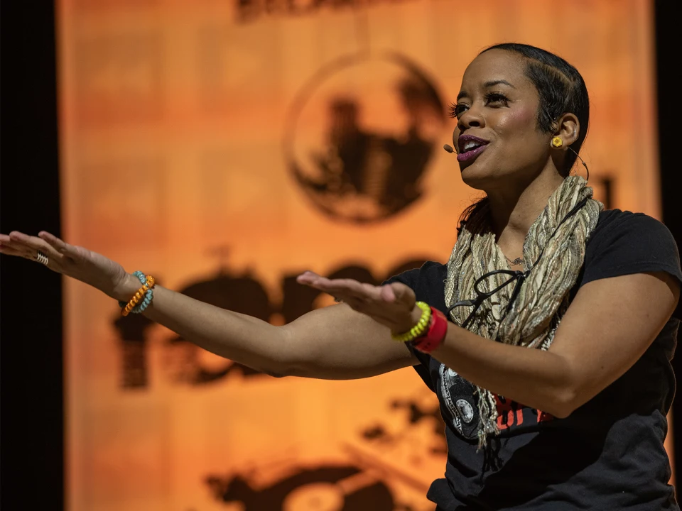 A woman wearing a scarf and bracelets extends her arms while speaking in front of an orange backdrop featuring black graphics and text.