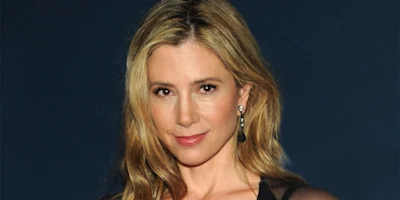 A woman with long blonde hair, wearing drop earrings and a black top, poses in front of a dark blue background.