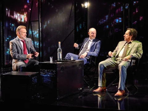 Three men in suits sit around a table with a bottle and glasses, against a backdrop of city lights at night.