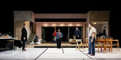 Five actors stand in a modern stage set resembling a living room and kitchen, with minimal furniture and props, engaged in what appears to be a dramatic scene.