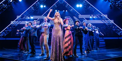 A group of performers, led by a woman in a gold dress, sing and gesture on a brightly lit stage with geometric light structures in the background.