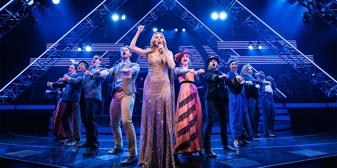 A group of performers, led by a woman in a gold dress, sing and gesture on a brightly lit stage with geometric light structures in the background.