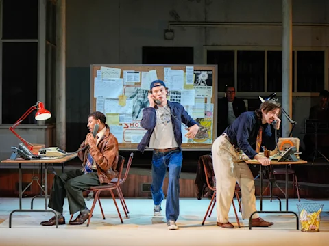 Three people in an office set, each using a phone at separate desks, with a bulletin board covered in papers and photos in the background.