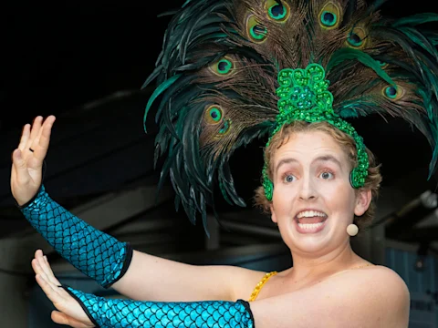 Person wearing a feathered headdress and blue sequined gloves performs with expressive gesture and wide-eyed facial expression.
