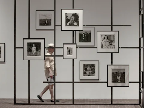 A man in light clothing and a hat walks past a grid-like display wall featuring multiple framed black-and-white photographs.