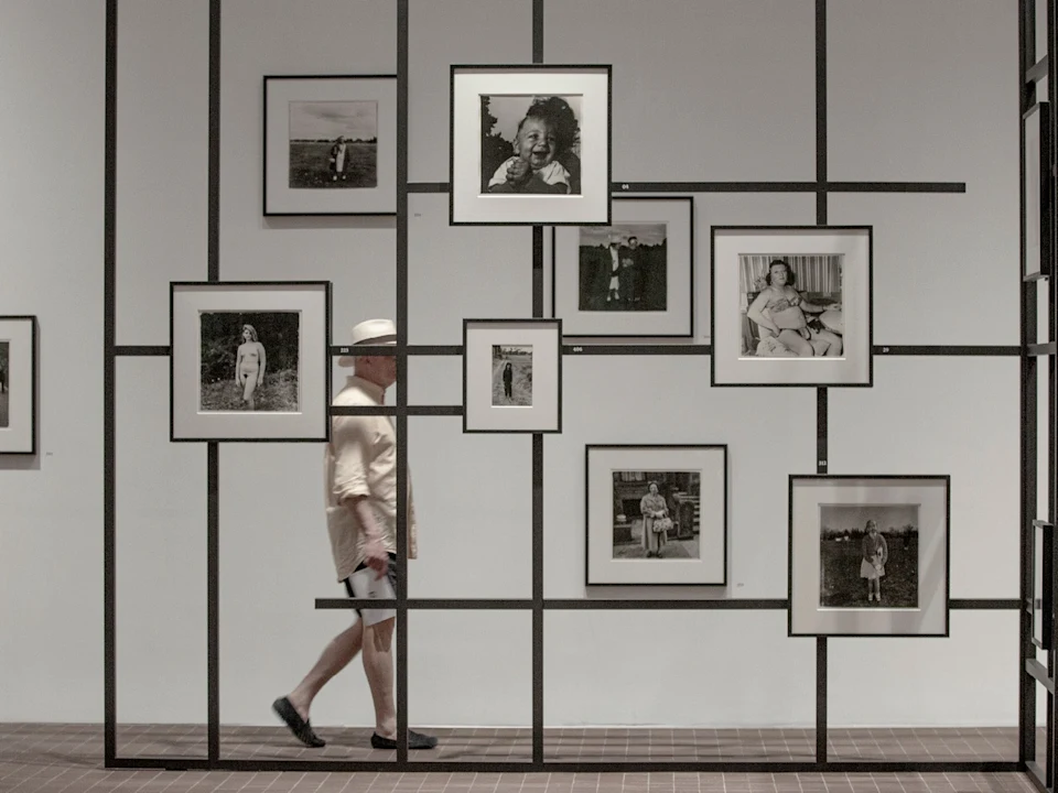 A man in light clothing and a hat walks past a grid-like display wall featuring multiple framed black-and-white photographs.