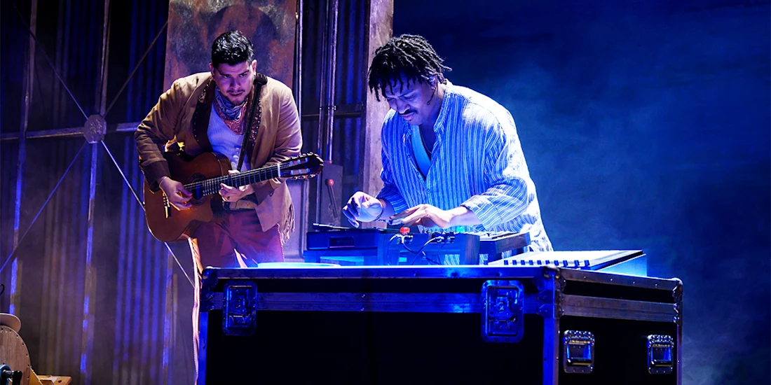 Two musicians perform on stage; one plays an acoustic guitar while the other operates a DJ controller on a lit equipment case.