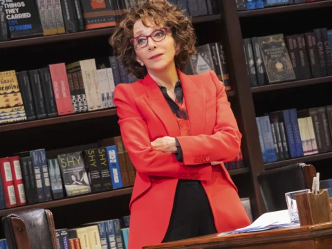 Person with curly hair and glasses in a red blazer stands with arms crossed in front of a bookshelf.