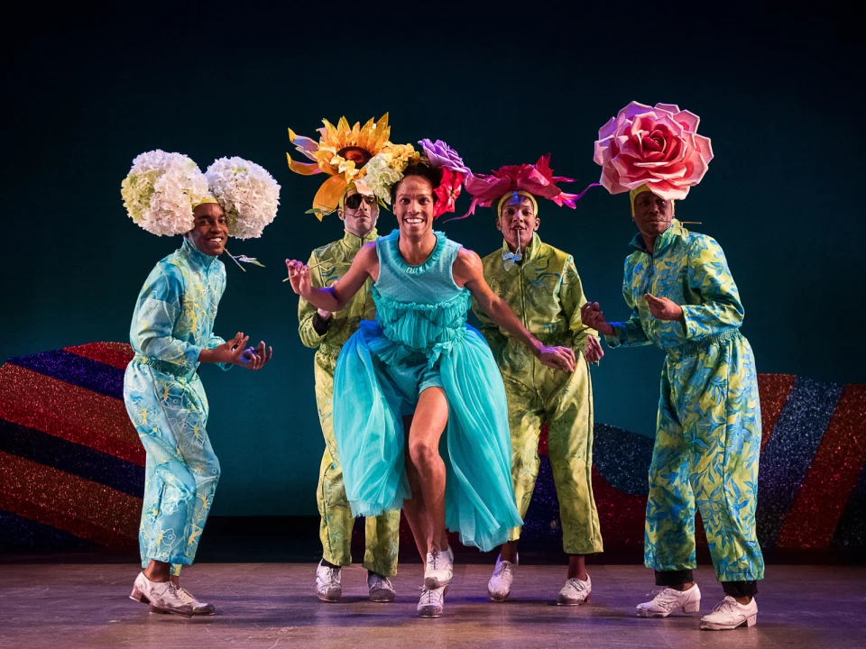 A group of five performers, four wearing floral headpieces and green floral costumes, and one in a blue dress, dancing on stage with a colorful backdrop.