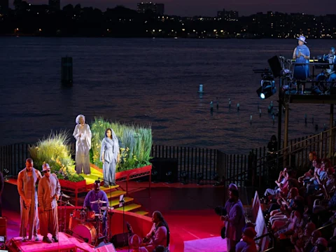 Actors perform on a brightly lit outdoor stage by the water at night, with musicians playing in front and an audience seated to the right.