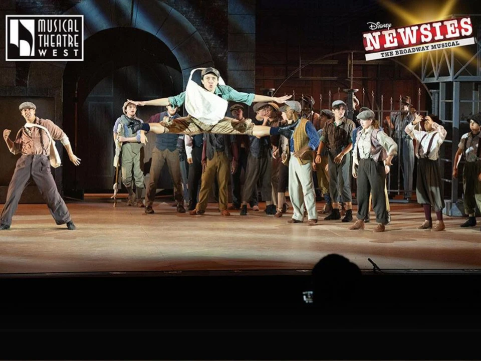 A stage performance of "Newsies" featuring dancers in mid-action. A jumping dancer is showcased prominently in the foreground, surrounded by other cast members in period costumes.