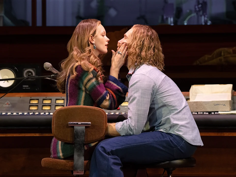 A woman gently touches a man's face as they sit close together in front of a recording studio console.