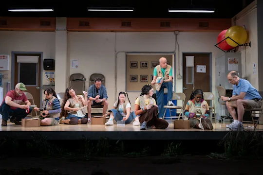 Nine adults sit and kneel on a stage set resembling a community center room, engaging in arts and crafts activities with focused expressions.