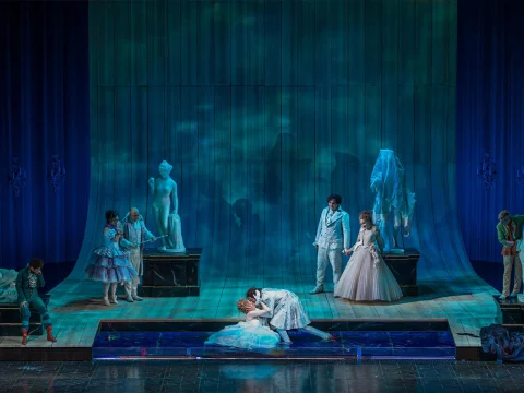 A theatrical performance depicts a dramatic scene with characters in elaborate costumes under blue lighting. Two central figures are locked in an embrace on a stage surrounded by others.