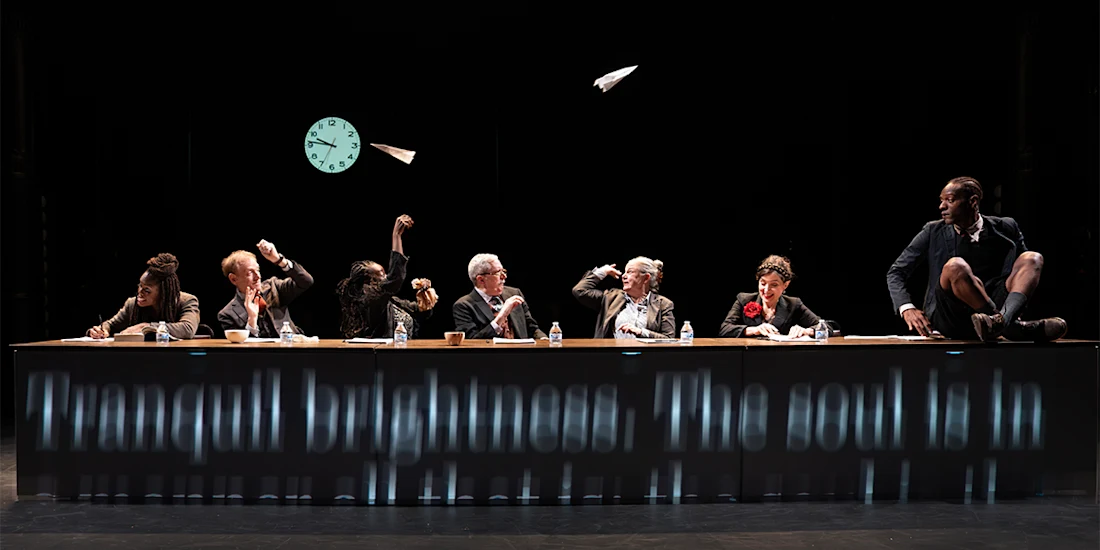 Seven people sit at a long table onstage, throwing paper airplanes; a large clock and a projected text are visible in the background.