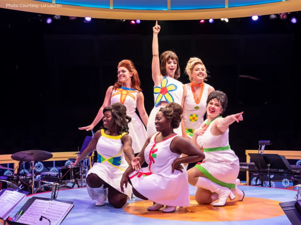 Six women wearing colorful dresses perform energetically on stage with musical instruments visible in the foreground.