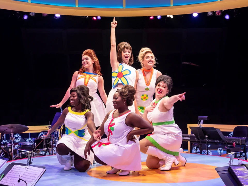 Six women wearing colorful dresses perform energetically on stage with musical instruments visible in the foreground.