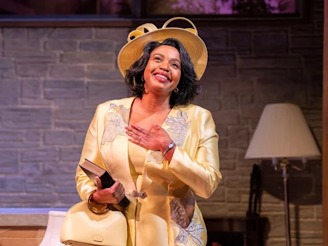 A woman in a yellow suit and hat smiles while holding a book and clutch, standing indoors near a lamp and stone wall.