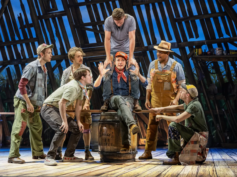 A group of men in colorful rural costumes gather around a man sitting on a barrel onstage, with a wooden barn set in the background.