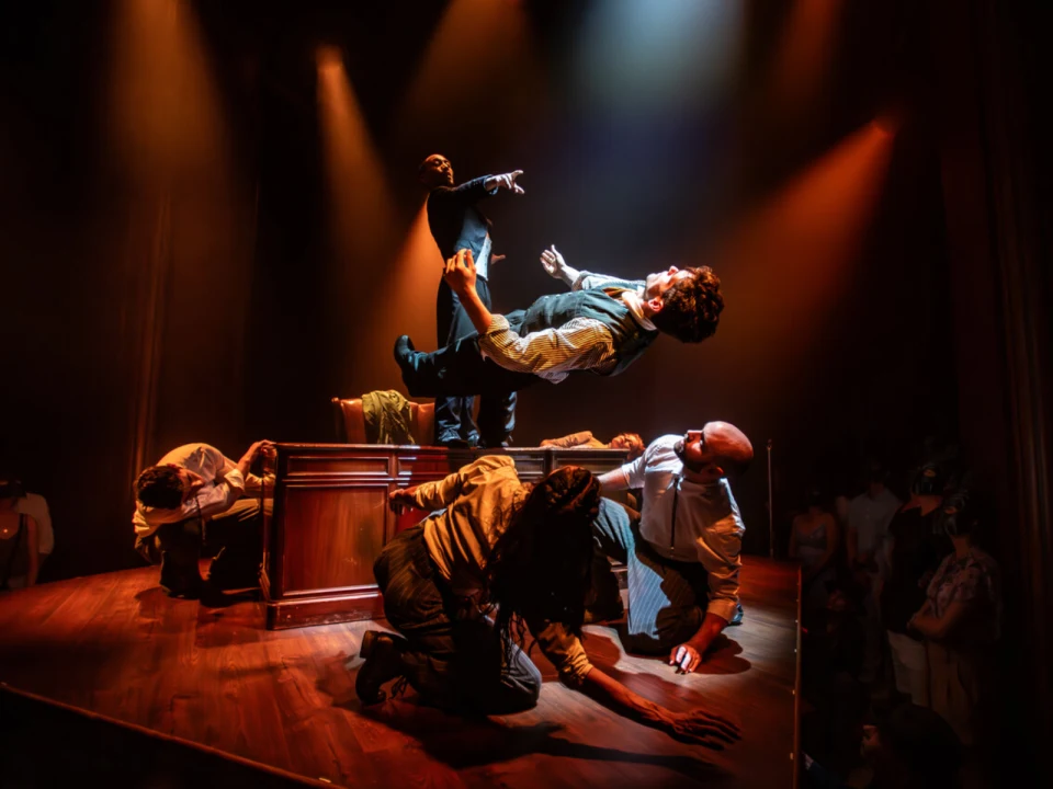 A group of performers in dramatic poses on stage, illuminated by warm lighting. One person stands on a table reaching upwards, while others kneel or crouch around them, creating a dynamic scene.