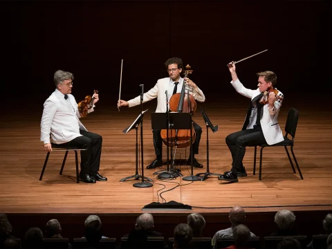 Three musicians perform on a wooden stage: a violinist, a cellist, and a violist, all dressed in white jackets and black pants. They are surrounded by music stands with an audience watching.