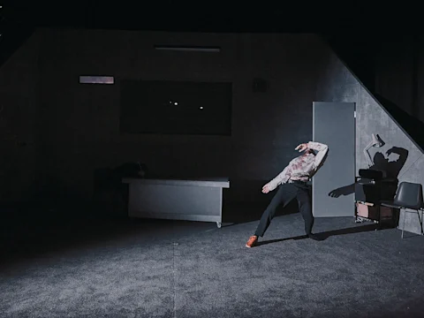 A person stands in a dramatic pose in a dimly lit room with a desk, chair, and open door; their shadow is cast on the wall behind them.