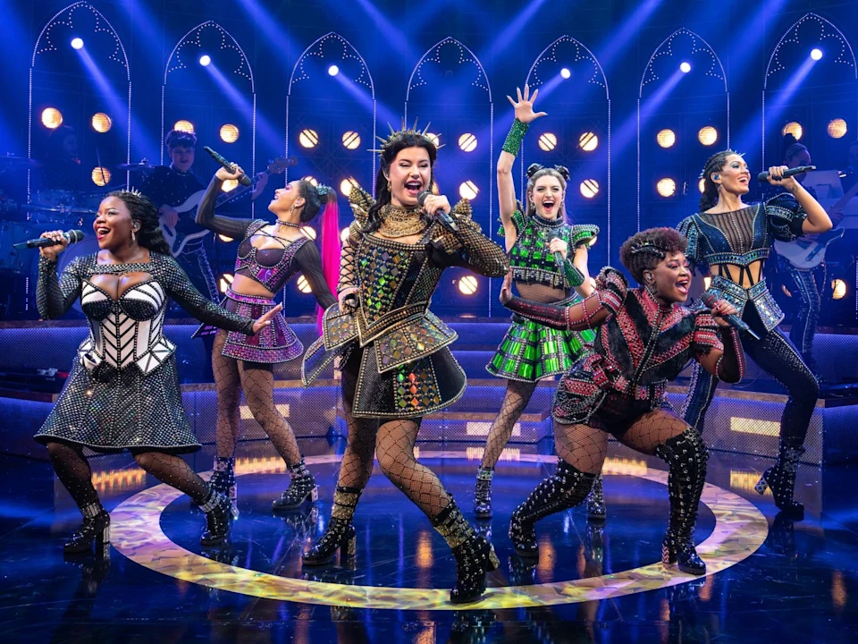 Production photo of SIX in Costa Mesa showing six women in elaborate, colorful costumes perform energetically on stage with microphones under dramatic blue and purple lighting.