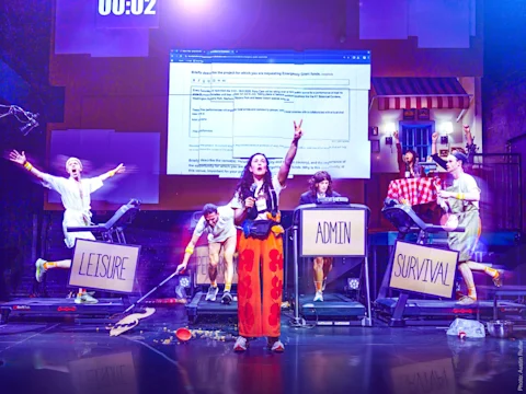 A stage scene with six actors, some on treadmills labeled "LEISURE," "ADMIN," and "SURVIVAL." One actor stands center stage, gesturing, with a large web page projected behind them.