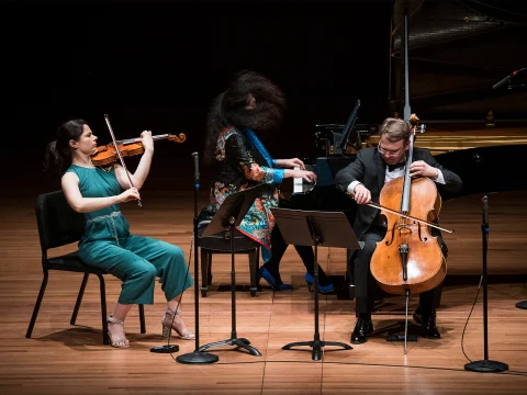 Three musicians perform on stage: a violinist in a green outfit, a pianist in a colorful dress, and a cellist in a black suit. They are seated, each playing their respective instrument.