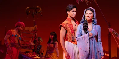 Actors in colorful costumes perform on stage in an Arabian-themed setting, with a man and woman standing together in the foreground and market stalls in the background.
