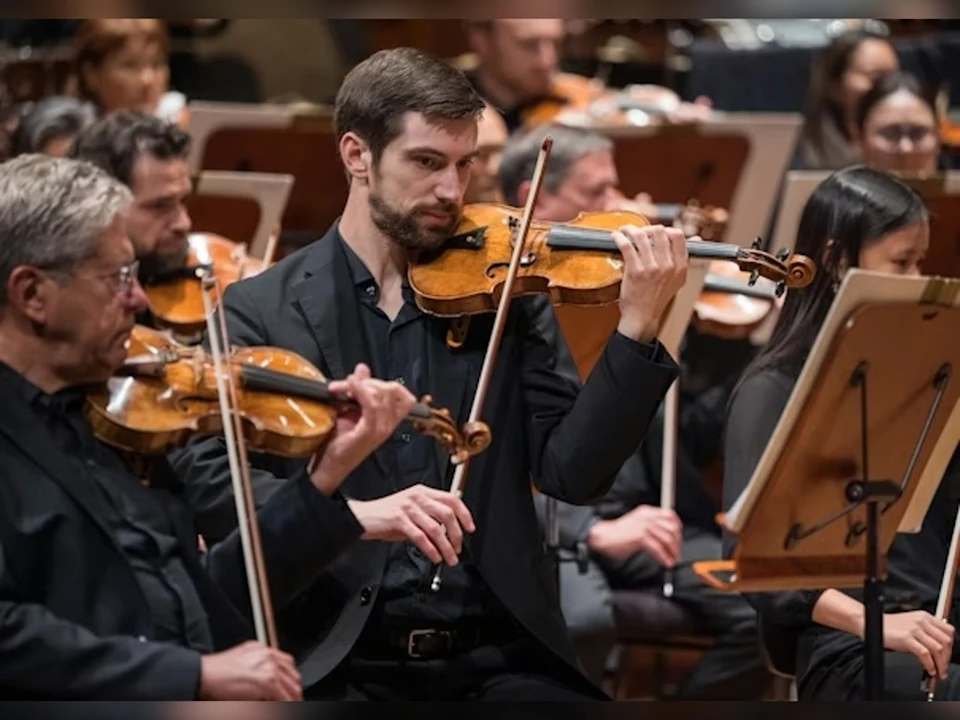 Several violinists in formal attire perform in an orchestra, focused on their sheet music and instruments in a concert setting.