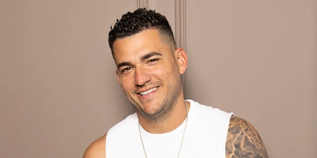 Man with short curly hair and a white sleeveless shirt smiles at the camera. He has a tattoo on his upper arm and stands against a plain beige wall.