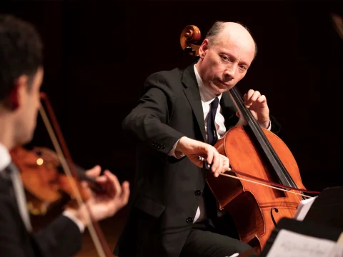 A man in formal attire plays the cello with focus on stage while another musician is visible in the foreground.