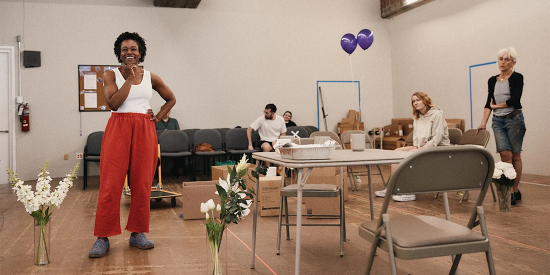 A woman in red pants stands smiling in a room with folding chairs, flowers, boxes, and people sitting and standing in the background.