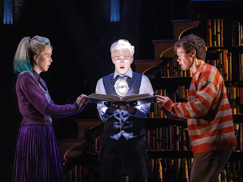 Three people stand in a library, holding an open book that glows with light. One person in the center wears glasses and holds a pocket watch above the book.